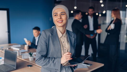 Muslim businesswoman in hijab looking at side working on digital tablet on colleagues background